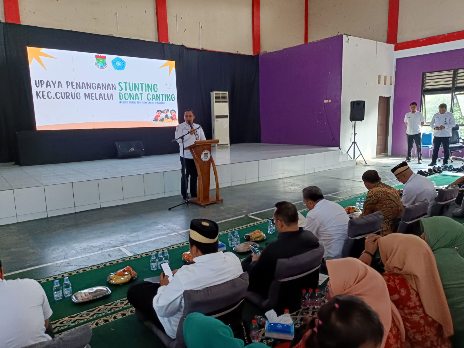 "Salah satu pejabat menyampaikan sambutan di atas panggung dalam kegiatan Program Donat Canting yang digelar di Gedung Serbaguna Kecamatan Curug, Kabupaten Tangerang, sebagai bagian dari upaya percepatan penanganan stunting. Acara dihadiri oleh unsur pemerintah daerah, TNI, dan tokoh masyarakat."